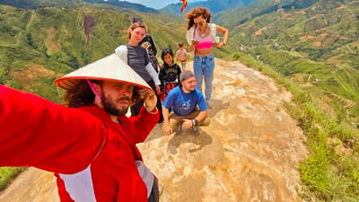 A group of six people are gathered on a dusty, uneven surface at the edge of a steep mountain landscape, possibly in Bắc Yên, Vietnam. The foreground is dominated by a man wearing a conical hat and a bright red jacket, holding up a camera to take a selfie. His expression is serious. To his right and slightly behind him, a woman with curly brown hair, wearing a pink crop top and blue jeans, is looking at her phone. A younger woman with blonde hair in a grey t-shirt stands next to her, also looking down. Two children, a girl in a black hoodie with "REGRESS" printed on it and a striped shirt, and a boy in a red and white striped shirt, are positioned in the middle ground. A man wearing a blue t-shirt and a baseball cap is crouched in the lower right quadrant of the image, looking to his right with a slight smile. The background reveals a vast valley with terraced green hills and winding roads, indicative of a mountainous rural area. The sky is bright and cloudy.