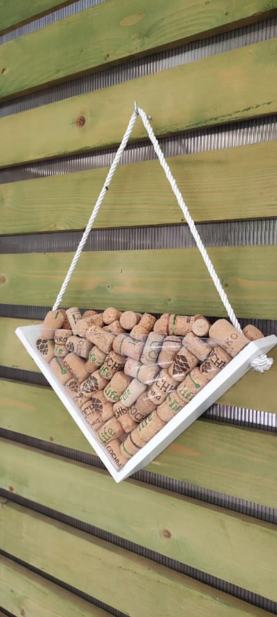 This media file shows a triangular white wooden shelf filled with wine corks, suspended by a white rope against a backdrop of weathered green wooden planks. The corks are varied, some with green lettering and designs, others plain. Some visible text on the corks includes "LIFE", "СОСНЫ", and what appears to be a logo with "KO". The shelf is hung against a textured, vertical gray background that runs between the wooden planks. The overall scene appears to be outdoors, possibly a porch or patio, given the wooden structure and the lighting. The image does not depict any people or animals, and there are no strong indicators of weather conditions other than ample light suggesting daytime. The setting seems to be a decorative display, possibly in a garden or outdoor living space. The location context is Dinskaya, Russia.