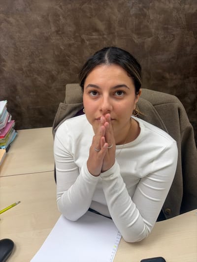 An individual, identified as a young adult female, is seated at a light-colored wooden desk in an indoor office environment located in Cagnes-sur-Mer, France. She has dark hair, parted centrally, and fair skin. She wears a white long-sleeved top, gold hoop earrings on both ears, and a thin gold chain necklace. Two silver-colored rings are visible on the thumb and index finger of her right hand. Her hands are clasped together, fingers interlocked and palms pressed, resting on the desk directly in front of her, beneath her face. She is looking directly towards the viewer.
On the left side of the desk, a stack of multi-colored papers or files is present, alongside a yellow pencil. A blank white sheet of paper, with faint markings along its right edge, lies directly in front of the individual. A dark object, possibly a computer mouse, is partially visible on the bottom right corner of the desk. A dark brown jacket or coat is draped over the back of the chair occupied by the individual. The background consists of a solid wall with a mottled, textured brown finish.