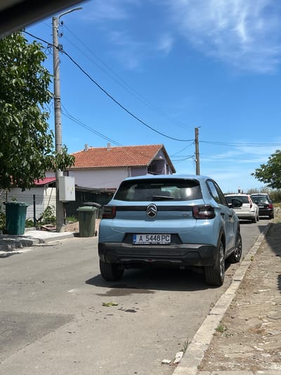 In Aheloy, Bulgaria, on a sunny day with a clear blue sky and scattered clouds, a light blue Citroen C4 is parked on an asphalt street next to a stone curb. The car, facing away from the camera, displays a license plate reading "BG A 5448 PC". Behind the Citroen, a white Volvo station wagon and a dark-colored sedan are also parked on the street. On the left side of the street, a green recycling bin stands near a fence and a building with a red tiled roof. Further down the street, another green bin is visible. A concrete utility pole with electrical wires runs along the side of the road. The environment appears to be a residential area with houses and vegetation.