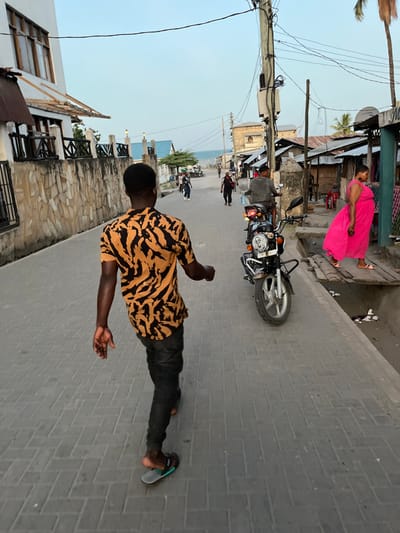 The photograph is rotated 90 degrees clockwise, depicting a lively street scene in what appears to be a coastal town, likely Bagamoyo, Tanzania.
In the foreground, centered, a man with dark skin walks away from the viewer, looking towards the right side of the frame. He wears a short-sleeved shirt with a distinctive black and orange tiger-stripe pattern and dark trousers. His left foot is bare, while his right foot is in a blue and white flip-flop with the word "AFRICA" visibly printed on its strap.
Behind him, on the same paved street made of grey interlocking bricks, a black motorcycle is parked. Further down the street on the left, a woman in a vibrant fuchsia-pink sleeveless dress stands near a building, facing left. Several other figures are visible in the distance, suggesting street activity.
The street is lined with buildings of varying construction. On the left, a low stone wall with wooden railings fronts a building with a white facade and dark wooden elements. On the right, older structures with corrugated metal roofs and stone or concrete walls are present. Tall utility poles with numerous tangled wires run along the right side of the street. Palm trees are scattered throughout the scene.
In the far background, under a clear, pale blue sky, the calm waters of the ocean are visible, suggesting the town is situated directly on the coast. The lighting indicates it is daytime, possibly late afternoon, with soft, even illumination. The scene conveys the atmosphere of a typical, bustling local street in a warm, coastal environment.