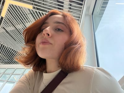 The image shows a person with red hair, light skin, and neutral-toned lips, positioned upside down. The person is wearing a light-colored top and a dark brown strap is visible across their shoulder. The background features a window and a grid-like structure with white and black lines, as well as a yellow light fixture. The location is potentially an interior space in Ianeti, Georgia, as requested.