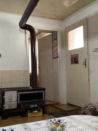This image depicts an interior residential space within the city of Branichevo, Bulgaria. The room features white painted walls and a light-colored ceiling. The floor is covered with brown tiles, over which two small, patterned dark rugs are placed near the center of the scene.
A dark, metallic wood-burning stove or cooker is positioned against the left wall, with two circular access doors on its left side and a larger rectangular oven door on its right side that is partially open. A light-colored wooden crate containing small wood pieces rests on the floor directly beneath the open oven door. Above the stove, a section of beige square tiles covers the wall. A dark brown, segmented metal stovepipe extends vertically from the stove, bends sharply to the right, and continues horizontally across the upper left corner of the frame.
Two doorways are visible in the background. The doorway behind the stovepipe is open, revealing a darker interior space with a faint glimpse of brown furniture. Two calendars are affixed to the wall or doorframe above this open doorway. To the right, a closed, light-colored wooden door features a frosted glass panel in its upper section and a metal handle. A small, colorful picture or calendar is affixed to this door below the glass panel. An electrical outlet with two sockets is mounted on the wall to the far right.
In the foreground, a white tablecloth with a patterned design covers a table, on which a clear glass bowl holds several individually wrapped candies. No individuals are present, and no active interactions are depicted.