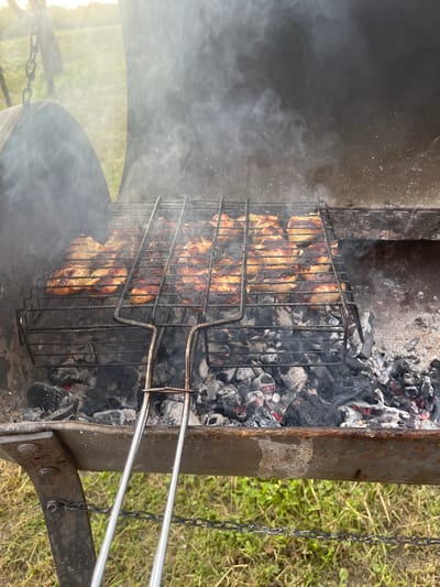 A close-up, eye-level, outdoor shot shows pieces of meat cooking on a grill over hot coals. The grill is black metal with a wire rack holding multiple rows of meat. The meat appears to be chicken pieces, some are well-cooked and browned, while others show signs of charring. Smoke rises from the grill, obscuring the background slightly. The grill itself is set within a larger, dark metal barbecue pit. Below the grill, glowing red coals are visible. The foreground features two silver metal grill tongs, one slightly out of focus. The background consists of green grass and a hazy, sunlit sky. The scene is consistent with a backyard barbecue setting, possibly in the city of Kalnciems, Latvia, given the context provided. There are no people visible in the image.