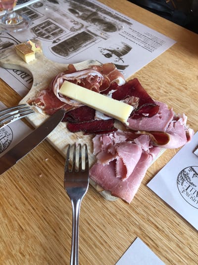 A top-down shot shows a wooden cutting board topped with various cured meats and cheese, alongside a fork and knife on a wooden table. The cutting board holds slices of light pink ham, dark red dried meat, and a rectangular piece of white cheese. A small cube of cheese rests near the edge of the board. A fork and knife are positioned across the cutting board. Adjacent to the board is a printed paper with a black and white architectural illustration and French text. Another paper with the words "AUBERGE DE LA" and "ALLE" printed on it sits on the table. The text references Gruyères. The scene appears to be in a restaurant or dining establishment, likely in Epagny, Switzerland.