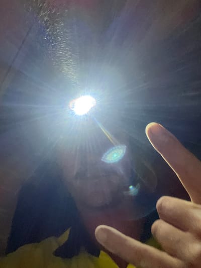 The image depicts a low-light indoor or enclosed environment in Wombarra, Australia, dominated by a single, intensely bright light source positioned in the upper center. This light emits strong radial flares and a distinct green/blue lens artifact below it. A person's face, obscured by shadow and the intense light, is partially visible in the lower-middle, with indistinct facial features. The individual appears to be wearing a yellow garment, possibly a jacket or shirt, visible at the bottom. A right human hand is prominently featured in the lower-right, performing a "rock and roll" or "horns" gesture, with the index finger and pinky extended and the other fingers curled. The background is dark and featureless beyond the immediate light.