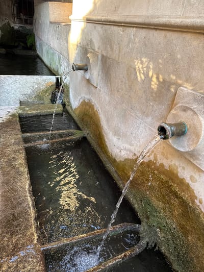 The image shows an outdoor public fountain in Moustiers-Sainte-Marie, France. Two spigots are mounted on a stone wall, with water flowing from them into tiered rectangular basins below. The stone is weathered and has patches of green algae and mineral deposits. The water is clear and reflects light. One basin appears to be overflowing into the next. The overall environment suggests a rustic and historic setting, typical of a Provençal village. There are no people visible in the image.