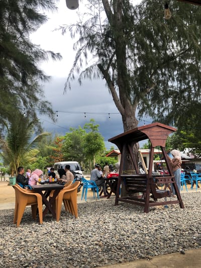 The image depicts an outdoor dining area at what appears to be a restaurant or cafe in Lancok, Indonesia, situated near a coastline indicated by the presence of palm trees and the type of vegetation. The ground is covered in gravel, and a large tree with pine-like needles dominates the center of the frame. A wooden swing set is positioned to the right, with a woman wearing a hijab and jeans standing near it, looking at her phone. Several tables and chairs, some made of brown plastic and others blue metal, are scattered throughout the area. A group of people, including adults and children, are seated at a table on the left, engaged in conversation and eating. They appear to be a family or group of friends. In the background, a white van is parked, and more greenery and buildings are visible. The sky is overcast and dark, suggesting either cloudy weather or impending rain. The overall atmosphere suggests a relaxed, casual dining experience in a tropical setting.