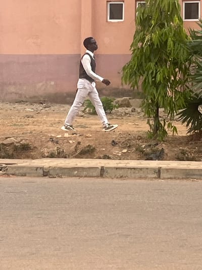 A person wearing a black vest over a white shirt and white trousers stands in an unpaved area adjacent to a building and a street in Badiko, Nigeria. The building is peach colored with square windows. A green plant grows near the person. The street is concrete. The ground is bare and littered with debris.