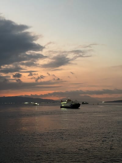 The image depicts a twilight scene over the ocean, likely near Melaya, Indonesia. Several ferries and smaller vessels are visible on the water, some illuminated by lights. The sky is painted with hues of orange and grey from the setting sun, with scattered clouds adding texture. The water is calm, reflecting the sky and the lights of the boats. In the distance, a silhouette of mountains or hills lines the horizon. The overall atmosphere is serene and captures the end of the day.