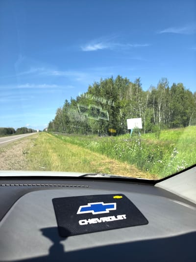 The image is a photograph taken from the interior of a vehicle, looking out through the windshield. The viewpoint is low, showing the dashboard in the foreground. On the dashboard, there is a black mat with the Chevrolet logo and name in white and blue. The windshield is slightly dusty and reflects the sky and the scene outside.

Outside the vehicle, a paved road stretches into the distance to the left. The road is bordered by a grassy shoulder and a field of green grass with scattered white wildflowers. To the right of the road, a dense line of trees, primarily deciduous with some birch, forms a forest. Some of the trees are in bloom or have white bark, characteristic of birch trees. Beyond the wildflowers and trees, a blank white sign is mounted on blue posts, and a smaller yellow sign is visible behind it. In the distance, a white truck can be seen on the road. The sky is clear blue with a few wispy white clouds. The overall environment appears to be rural or a highway passing through a natural landscape.

The specific location mentioned is "Пригородный ауылдық округі, Kazakhstan." This suggests the scene is in a rural district or settlement near a city in Kazakhstan. The environment depicted, with a highway, fields, and a forest, is consistent with a roadside view in a region that could be near such a settlement.
