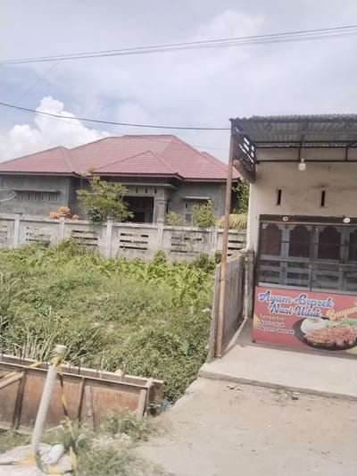 A sunlit outdoor scene depicts a residential building with a red tiled roof in the background, flanked by a concrete fence. In the foreground, a building with a corrugated metal roof overhang houses a food stall. A banner on the stall advertises "Ayam Geprek Nasi Uduk Bayu," indicating the availability of fried chicken with coconut rice and a signature dish. The surrounding environment is overgrown with lush green vegetation. The image appears to be taken in the city of Beurandang, Indonesia, suggesting a street view of a local eatery and its surroundings.