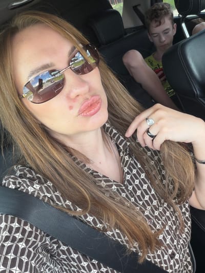 A woman with long, brown hair, wearing sunglasses and a brown and white patterned shirt, is taking a selfie in the front passenger seat of a car. Her lips are pursed, and she is wearing rings on her fingers. In the back seat, a young boy with curly brown hair is looking down at his phone. The interior of the car is visible, including black leather seats and a seatbelt. The image appears to be taken during daylight, with sunlight reflecting off the sunglasses. The location in the city of Severny, Russia, is not discernible from the image.