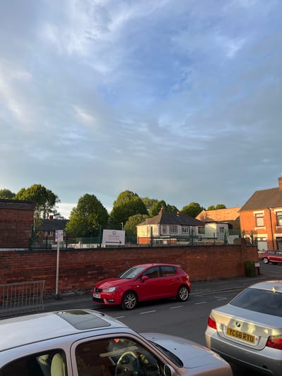 The image depicts a street scene in Stoke-on-Trent, United Kingdom, with a focus on parked vehicles and the facade of buildings. A red hatchback, possibly a SEAT Ibiza due to its license plate "SK55 LXH", is parked on the right side of the road. To its left, a silver BMW sedan with license plate "YC58 RTO" is also parked. In the foreground, a silver car, partially visible, has its sunroof open. The background reveals a brick wall and a fence surrounding buildings, including what appears to be an educational institution with a sign that reads "EXCELLENCE GIRLS ACADEMY". The sky above is filled with a mix of clouds and clear blue patches. The lighting suggests it is either early morning or late afternoon, casting long shadows.