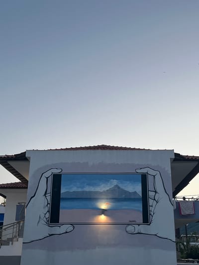 The image shows a mural on the side of a building in Toroni, Greece, depicting a smartphone held by two hands, displaying a scene of a beach with a mountain in the background. The sky is clear and pale blue, with a hint of sunset glow. The building has a tiled roof and white walls. There is a staircase with a metal railing leading up to the building on the left side. On the right side of the building, a balcony with some clothes hanging on a line is visible. The mural is rendered in a realistic style, with the hands and the image on the screen appearing three-dimensional. The mural is signed by "KARABABAS".