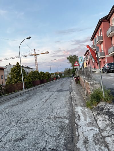 This image captures a quiet street scene in Camerino, Italy, during what appears to be late afternoon or early evening, characterized by a soft sky with pale blue hues and faint pink clouds. The view looks down a straight, asphalt road that exhibits significant cracking and wear.

On the left side of the street, a row of multi-story residential buildings, primarily painted in reddish-pink tones, line the thoroughfare. These structures feature multiple windows, some with external shutters or blinds, and balconies. A grey Dacia Duster SUV is parked by the curb in front of one of these buildings; its license plate is partially visible, starting with "CW 071 Y".

The right side of the road features a cracked concrete curb or sidewalk, with grass and weeds growing through its fissures. Several modern streetlights arc over the road. Prominent road signs are visible on the left: a red triangular warning sign with a black zigzag symbol indicating a winding road, and below it, a white rectangular sign displaying "90 m". Another blue rectangular sign with white pictograms, possibly indicating a pedestrian or cycling path, stands nearby.

Further into the distance, trees and green hedges flank the road, partially obscuring other buildings. A large, yellow lattice construction crane is clearly visible against the sky in the background, suggesting ongoing development. The scene is devoid of people, contributing to its serene and calm ambiance.