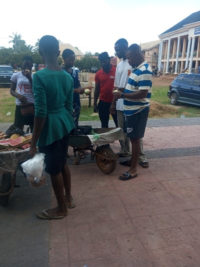 The image displays an outdoor scene in Ajalli, Nigeria, depicting individuals gathered around a street vendor setup on a paved surface. In the foreground, a person wearing a green peplum top and dark skirt stands with their back to the viewer, holding a clear plastic bag containing items.
To their left, a cart displays sliced watermelon and other produce. Adjacent to this, a green wheelbarrow or second cart is present, containing items partially covered by a white bag. Six individuals are visible in the mid-ground, interacting near these vendor stations. From left to right: a female in a white t-shirt consumes a light-colored fruit; a female in a patterned blue dress holds a similar fruit; a male in a red t-shirt stands; a male in a white t-shirt stands; and a male in a blue and white striped t-shirt holds a light-colored fruit.
The immediate ground is covered by patterned red paving, transitioning to a grassy and dirt area further back. Concrete blocks are stacked near the vendor area. In the background, a large multi-story building with prominent white columns and a blue roof is visible, bearing partially legible signage that reads "HOLY...". Parked vehicles include a dark SUV on the left and a blue hatchback on the right. The sky is overcast.