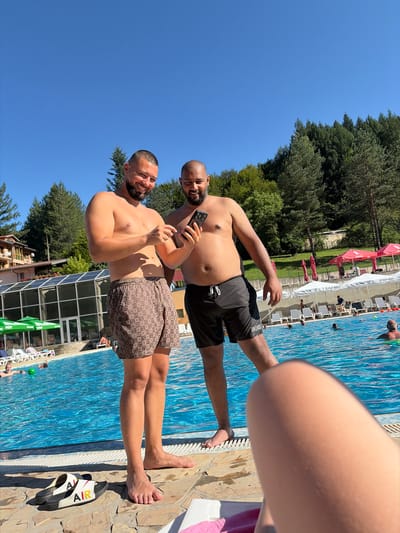 Two adult males, appearing to be in their 30s or 40s, are standing by the edge of a bright blue swimming pool. The man on the left is wearing patterned swim trunks and is smiling while looking at a smartphone held by the man on the right. The man on the right is wearing black swim trunks and is also looking at the smartphone. Both men are bare-chested. The background shows a resort with a building, trees, and more swimming pool area with lounge chairs and umbrellas. The scene is outdoors under a clear blue sky. This is likely the resort located in Chiflik, Bulgaria.