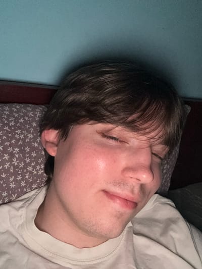 The image shows a close-up, head-and-shoulders shot of a young man with brown hair and closed eyes, resting his head on a patterned pillow. He is wearing a white t-shirt. The background consists of a red headboard and a turquoise wall. There is no indication of the location being in the city of Dnipro, Ukraine.