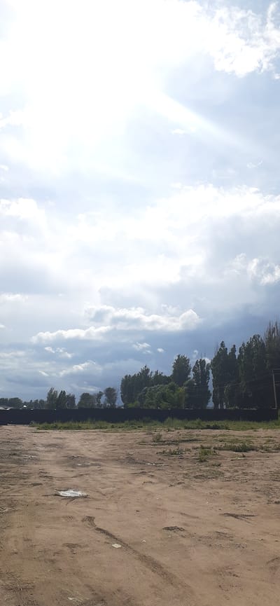 The image depicts an outdoor scene on a partly cloudy day, likely in Chok Tal, Kyrgyzstan. The foreground consists of a dirt ground with tire tracks, suggesting it may be a pathway or an undeveloped area. In the midground, a black fence runs horizontally across the image, separating the foreground from a strip of green grass and vegetation. Behind the fence, a line of trees, varying in height and density, forms a natural barrier. The sky occupies the upper portion of the image, with large, white clouds and streaks of sunlight indicating a bright, though not entirely clear, day. There are no people or animals visible in the frame.