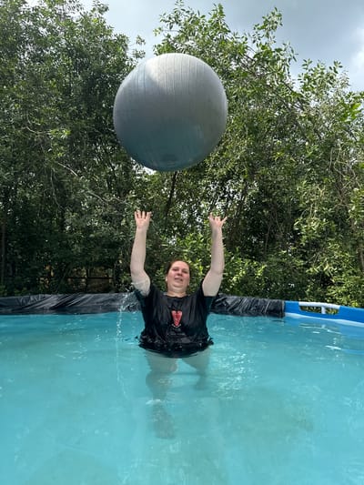 A person is standing in a blue swimming pool filled with clear water. They are reaching up with both hands towards a large, grey exercise ball suspended in the air above them. The person is wearing a black t-shirt that is wet and clinging to them, with a red emblem on the left chest. Water is splashing around them. The pool is surrounded by lush green trees and foliage, indicating an outdoor setting. The sky above is partly cloudy. The overall scene appears to be a recreational activity taking place in a backyard or a natural outdoor area. The location is identified as Khokhryaki, Russia.
