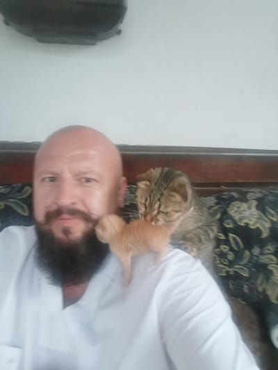 In this selfie, a bald man with a mustache and beard is pictured from the chest up. He is wearing a white shirt. He is looking directly at the camera with a neutral expression. To his right, a tabby cat is perched on his shoulder, nuzzling a small orange kitten. The kitten is positioned on the man's left shoulder, facing away from the camera. The background shows a dark wooden headboard of a bed with a patterned dark fabric on the pillows. The lighting suggests it is daytime, and the setting appears to be indoors, likely a bedroom. The overall mood is relaxed and affectionate, highlighting the bond between the man and his pets. There is no visible text in the image. The location context provided is Semypolky, Ukraine.
