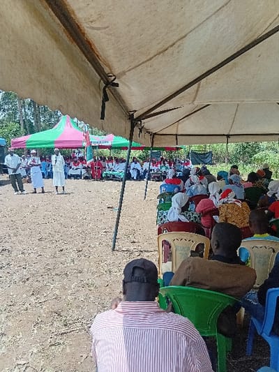 The image captures an outdoor public gathering in Butali Chegulo ward, Kenya, under a bright sky. In the foreground, a large, light-colored tent provides shade, under which numerous individuals are seated on various plastic chairs (beige, green, blue) on a dry, light-brown, dusty ground. The backs of several attendees are visible, including a male in a red and white striped shirt and a dark cap positioned prominently in the immediate foreground.

Further back, a second large tent with distinct green and pink stripes shelters additional participants. Within this tent, on the left, two individuals stand; one, clad in a long white garment and a head covering, faces the seated audience, appearing to address them. To their right, several persons are seated at tables, many wearing white robes with red sashes or stoles. A banner with visible text is positioned behind this seated group. Scattered trees and sparse shrubbery form the distant background. A speaker or sound system component is partially visible on the far left.