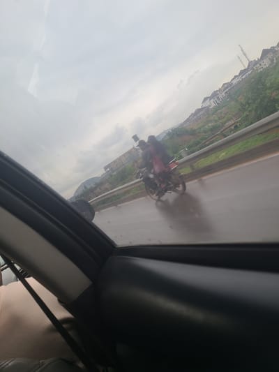 In Kugbo, Nigeria, a motorcycle with two passengers travels on a wet road under an overcast sky. The foreground shows the interior of a vehicle, with a black car door frame and window visible. The middle ground features the highway with a metal guardrail separating it from a grassy embankment dotted with houses. In the distance, more houses and communication towers are visible on a hillside. The motorcycle, with a rider and a passenger in the back, is captured in motion, casting a reflection on the wet asphalt. The overall scene suggests a journey through a suburban or semi-urban area on a cloudy day.