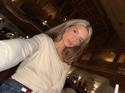 A woman with blonde hair, light skin, and light blue eyes poses indoors. She wears a cream-colored long-sleeve shirt and blue jeans with a Tommy Hilfiger patch. The background features a rustic interior with exposed wooden beams and dim lighting. Tables and chairs are visible in the distance, suggesting a restaurant or lodge setting.