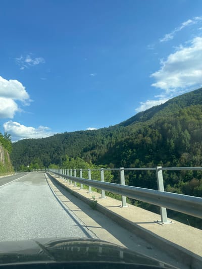 The image depicts a scenic view from a car driving on a road that curves to the left, ascending a mountainside covered in dense green forest. The road is paved with asphalt and has a concrete barrier with a metal railing on the right side. The sky is a clear blue with scattered white clouds. The sun casts long shadows from the railing onto the road and the concrete barrier. The location is on the road leading to the town of Taran, Bulgaria.