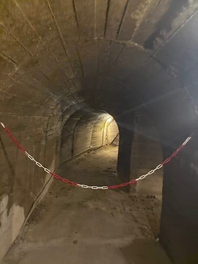 The image displays a segment of an underground tunnel identified as part of the Znojmo Underground in Znojmo, Czechia. The tunnel possesses an arched ceiling and walls constructed from rough, light-colored stone or concrete, exhibiting visible texture and signs of dampness. An alternating red and white plastic chain spans horizontally across the tunnel in the mid-ground, functioning as a physical barrier. The chain presents a slight sag in its center. The tunnel floor consists of an uneven, packed earth surface. A thin, dark cable is routed along the lower section of the left wall. Artificial light emanates from a source positioned deeper within the tunnel, illuminating the distant section and casting a forward glow. No individuals, actions, or interactions are visible within the scene.