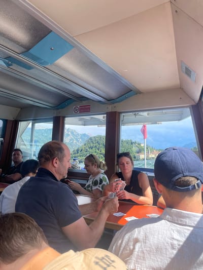 People are playing cards at a table with a view from the window. The outside shows a landscape with water, mountains, and a building with a red flag. There are several other people partially visible around the table. The inside is light with paneled walls and windows with a sign in Italian. The overall image is somewhat tilted.