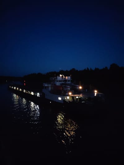 A large barge is illuminated at night on a body of water in Uzhla, Russia. The barge's structure includes a taller section towards the front, likely housing the bridge and crew quarters, and a long, lower cargo area trailing behind it. Several lights shine from the barge, reflecting in the water. Trees line the shore in the background, silhouetted against the night sky.