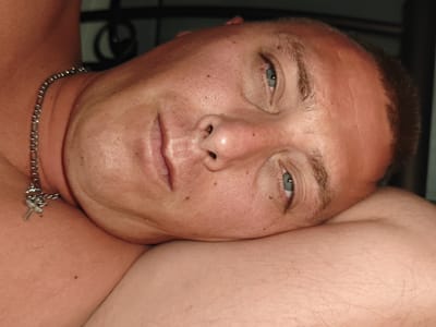A close-up, indoor shot depicts a person with fair skin, short brown hair, and blue eyes. They are lying down with their face resting on their arm. A silver chain necklace with a cross pendant is visible on their chest. The background is dark and slightly out of focus, revealing an indistinguishable dark object.

There is no information present in the image to determine if the location is in Gouves Municipal Unit, Greece.