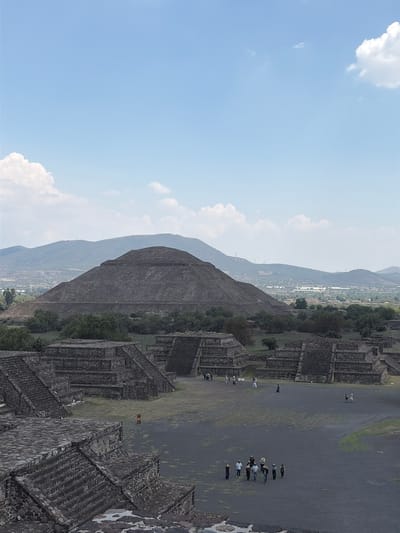 The image presents an angled view of the Teotihuacan archaeological site in San Francisco Mazapa, Mexico. The Pyramid of the Sun dominates the scene, flanked by smaller structures and mountainous terrain in the background. A wide, grey, paved expanse is visible, featuring scattered groups of people. The sky is predominantly blue with scattered clouds. The architecture of the pyramid and the surrounding structures is composed of dark stone blocks. The environment appears arid and dry.