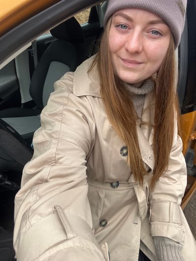A young woman with fair skin and light brown hair is seated inside a vehicle, possibly a car, with an orange door frame visible on the left. She wears a beige trench coat over a grey sweater and a grey beanie. The interior of the vehicle features dark seats with light grey inserts. The photograph appears to be a selfie taken by the woman. There is no evidence in the image to suggest it was taken in Ivlevo, Russia.