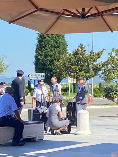 This media file captures an outdoor scene in Golubovci, Montenegro, likely during daytime with clear skies and sunny weather. The foreground features a large, partially visible beige umbrella, casting a shadow. A group of people, some in uniforms that appear to be airline crew or similar professional attire, are present.

One individual, wearing a dark uniform with insignia and a cap, is standing and looking towards the left. Another person in a light-colored uniform with a hat is also visible, facing a sign. Several other individuals in various uniforms and casual clothing are scattered throughout the frame. Some are standing, while others are seated or leaning. Luggage is visible near some individuals, suggesting travel or a departure/arrival point.

A prominent blue and white directional sign is visible, with text that reads "ČASOVNO HOURLY" and a parking symbol. This indicates a nearby parking facility and suggests a time-based service. The background shows lush green trees, foliage, and some buildings or structures, along with a road or paved area. The overall atmosphere appears to be one of an organized gathering or event, possibly at an airport or a similar transport hub, given the luggage and uniforms. The sun is bright, indicated by the shadows and the clear blue sky.