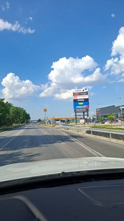 This is a first-person perspective, eye-level, medium shot taken from inside a vehicle, looking forward on a two-lane asphalt road. The road is lined with a metal guardrail on the right and trees on the left. In the distance, there are several commercial buildings and a tall billboard advertising various businesses. A Shell gas station with yellow pumps is visible to the left of the billboard. The sky is bright blue with scattered white cumulus clouds. The ground is paved and there are white lane markings on the road. The overall scene suggests a commercial area on the outskirts of a city, likely a highway exit. The text on the billboard indicates the location is in Skutare, Bulgaria, listing businesses such as "EAST\u00aeROAD", "BBQ HOUSE", "Sinsay", "TEDI", "pepco", and "MAGELAN CERAMICA".