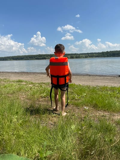 This media file shows a young boy wearing an orange life jacket, standing on the grassy bank of a body of water. The boy is facing away from the camera, looking towards the water and a tree-lined distant shore. The water appears calm, reflecting the bright blue sky filled with white, puffy clouds. The sun is high in the sky, suggesting it is daytime, likely midday or early afternoon. The weather appears to be warm and sunny.

The foreground is dominated by green grass and a sandy/dirt path leading down to the water. The boy's legs are visible, one bent at the knee, and he appears to be reaching out with his left arm. The overall scene conveys a sense of outdoor recreation and summer enjoyment. The location is identified as Pravda, Russia, which provides geographical context to the scene. There is no visible text in the image.