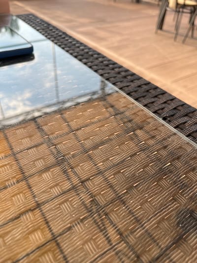 The image shows a close-up, angled view of a woven wicker pattern surface, likely a table or chair, covered by a clear glass surface. The glass reflects the sky and some greenery, suggesting an outdoor setting. The wicker pattern is a dominant element, with its intricate weave and earthy brown tones. A dark object, possibly a phone or a book, rests on the glass surface in the lower left corner, partially obscuring the reflection. The scene is brightly lit, indicating daylight. The overall impression is one of a casual outdoor setting, perhaps a patio or garden. The location context of Azov, Russia, does not provide any specific visual cues within the image itself. No text is visible.
