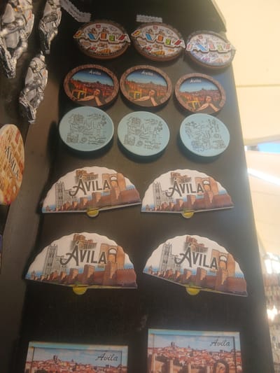 A collection of souvenirs, primarily magnets and possibly small plaques, are displayed on a dark, textured surface, likely a refrigerator door or a similar magnetic surface. The items predominantly feature imagery of Ávila, Spain, a historic city renowned for its well-preserved medieval walls.
The souvenirs include several round magnets with cityscapes and text. Some are circular with a wooden-like frame and depict Ávila's skyline with an indication of the city walls and buildings, along with text that appears to be "Ávila". Other circular magnets are flatter and lighter in color, featuring line drawings of architectural landmarks and streets within Ávila, again with the name "Ávila" prominently displayed.
There are also fan-shaped magnets, each depicting a stylized representation of the Ávila city walls and a cathedral or large church structure. The "Ávila" text is overlaid on these designs. One fan-shaped magnet is partially visible on the left side of the image, appearing to be a rectangular magnet with a circular image and text.
In the bottom portion of the image, two rectangular magnets are visible. The lower one shows a panoramic view of Ávila's rooftops and walls under a blue sky, with the word "Ávila" written in cursive. A portrait of a person, possibly a historical figure from the region, is partially visible in the lower right corner of this magnet.
The overall presentation suggests a retail display or a personal collection of mementos from Ávila, Spain, highlighting its iconic fortifications and architectural heritage. The lighting in the image is somewhat diffused, with a bright light source likely coming from the right, creating a slight glare on some surfaces.