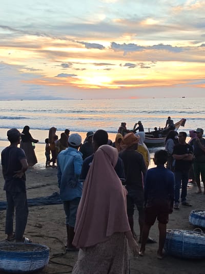 This image depicts a beach scene at sunset in Uleekareung, Indonesia. A group of people are gathered on the sandy shore near the water. Several individuals are standing, some facing the ocean and others interacting with each other. A traditional fishing boat is visible in the shallow water, with people around it, possibly preparing to launch or having just returned. Two large woven baskets, appearing to contain fish, are placed on the sand in the foreground. The sky is filled with a gradient of colors, ranging from pale blue to vibrant orange and yellow, indicative of a sunset. The ocean is calm, with small waves lapping the shore. The overall atmosphere suggests a community engaged in fishing activities at the end of the day.