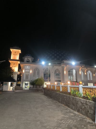 An exterior nighttime shot captures a mosque in Paya Tringgadeng, Indonesia. The mosque, illuminated by artificial lights, features a prominent blue-tiled dome and a tall, white minaret. Ornate windows and decorative banners adorn the building's facade. A paved walkway leads to the mosque, flanked by a low, stone wall with landscaping. The sky above is dark and clear.
