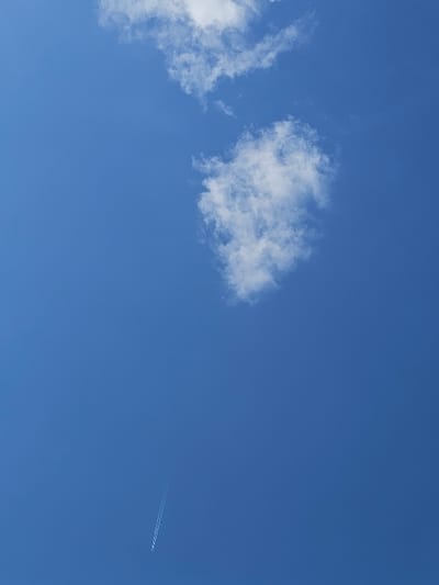 The scene depicts a clear blue sky above Donchevo, Bulgaria, under daylight conditions. Multiple white cumulus clouds are scattered across the upper half of the frame, with a larger cluster visible towards the upper right. A distinct, linear white contrail, characteristic of a high-altitude aircraft, originates from the lower-left corner and extends diagonally upwards and to the right, fading into the blue. No individuals, terrestrial objects, or specific interactions are visible within the frame, which focuses exclusively on the sky.