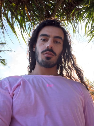 A low-angle, close-up, selfie-style portrait of a person with long, dark, wavy hair and a beard, wearing a pink t-shirt. The person is outdoors, with palm fronds and a bright sky visible in the background. The lighting suggests it is daytime, with sunlight filtering through the foliage. The scene appears to be in Selemadeg Barat, Indonesia, with lush vegetation typical of the region. The person is looking directly at the camera.