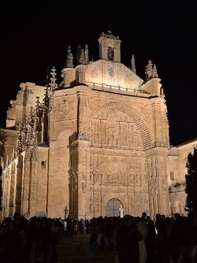 The image displays the illuminated Plateresque facade of the Convento de San Esteban (Dominican Monastery) in Salamanca, Spain, under a dark night sky. The large sandstone building features an intricately carved multi-tiered facade with numerous relief sculptures depicting religious figures, scenes, and heraldic emblems. A prominent arched entrance, fitted with dark iron gates, is located at the ground level, with a small, light-colored statue positioned directly in front of it. The upper sections of the facade include architectural elements such as columns, niches, pinnacles, and a visible bell in a belfry towards the apex. Artificial lighting highlights the building's details against the dark environment. In the foreground and midground, a dense crowd of approximately 50-70 individuals occupies the paved plaza before the monastery. These individuals are largely visible as dark silhouettes, standing and facing towards the illuminated structure. A dark tree or large shrub is partially visible on the far right of the scene.