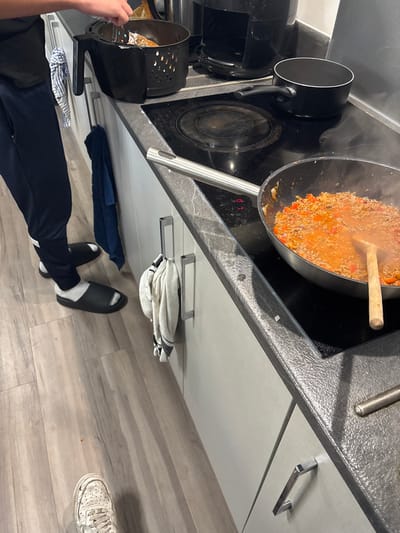 The image shows a kitchen counter with a black electric stovetop and a stainless steel pan on the right burner. The pan contains what appears to be a meat sauce with red peppers and is being stirred with a wooden spoon. The left burner is not in use. A black air fryer is on the counter to the left of the stovetop. A person's legs and feet are visible on the left side of the image. The person is wearing dark pants and white socks, and they are wearing two different pairs of shoes: black slippers on one foot and a white sneaker on the other foot. There are two towels hanging on the cabinet doors to the left of the stovetop, one blue and one white. The environment appears to be a kitchen in a domestic residence. The coordinates provided (latitude: 54.005, longitude: -2.79) place the location in Lancaster, United Kingdom.