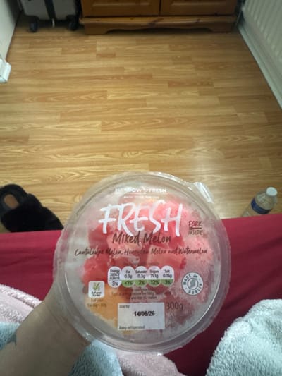 A close-up, slightly angled medium shot captures a clear plastic container of "Fresh Mixed Melon" fruit salad resting on a red surface, likely a bed or sofa. The container is approximately three-quarters full of diced red and orange melon pieces, with the "Fresh" logo prominently displayed in white text. A nutritional information label is visible on the container, showing values for energy, fat, saturates, and sugars per 100g and as a percentage of daily intake, along with a "Use by" date of 14/06/26. The words "Cantaloupe Melon, Honey Melon and Watermelon" are also printed on the label, and a "Made in Dublin" circular sticker is present. To the left of the container, a black slipper with a tan sole is partially visible. In the background, a light brown wooden floor extends horizontally. To the far right, a portion of a white radiator and a clear plastic water bottle with a blue label and white cap can be seen. The overall lighting is moderate, with some reflections on the plastic container. The image does not provide any specific information that would allow for the identification of the city of Armagh, United Kingdom.