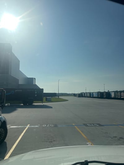 The image depicts an outdoor scene at what appears to be a logistics or distribution center. The sun is shining brightly, suggesting it is daytime, possibly morning or afternoon, with a clear blue sky. The setting is an asphalt lot marked with yellow and blue lines, indicating parking or loading bays.
Prominently featured are several large shipping containers, many bearing the Amazon Prime logo and branding. Some containers are stacked, while others are on the ground. One visible container has text that reads "STACK: UNIT 3 HIGH" and "TOP LIFT ONLY". Another container has "prime" and a blue Amazon logo. There are also what appear to be semi-trucks or tractor-trailers parked in a line. Some of these vehicles are white, and one in the foreground is partially visible and appears to be white as well.
In the background, a large, modern building with a reflective facade is visible, likely the distribution center itself. The lot is also lined with light poles. A patch of green grass with some bushes is visible in the middle ground. The ground is concrete or asphalt, with some scattered debris and water marks. There are numbers painted on the ground, such as "578" and "579", likely identifying parking spots or bays. The overall impression is one of a busy industrial area focused on shipping and logistics. No people are visible in the image.
