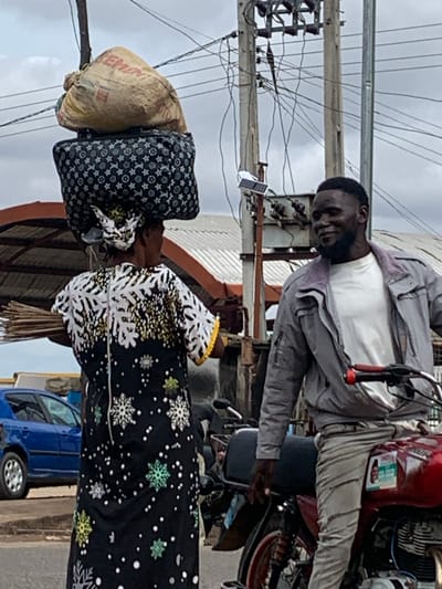 This image depicts an outdoor street scene in Shendam, Nigeria, under an overcast sky.
In the left foreground, a woman is observed from behind, carrying a substantial load balanced on her head. The load comprises a dark-colored, rectangular bag patterned with light-colored stars and symbols, surmounted by a light brown sack prominently labeled "CEMENT." She wears a long, dark-colored dress adorned with white and yellow snowflake-like patterns on the upper half and smaller, multicolored snowflake patterns with white dots on the lower half. A bundle of brooms is held in her left hand, extending horizontally.
In the right foreground, a man is seated on a red motorcycle with a black seat. He is facing partially towards the woman, exhibiting a mild smile or pleasant expression. He has short dark hair and a beard, and is dressed in a white t-shirt, a grey zippered jacket with a visible purplish collar lining, and light-colored trousers.
The background features several utility poles supporting a complex network of electrical wires against the grey sky. A small solar panel device with an attached light is mounted on one of the poles. To the far left, a blue car, identified as a Peugeot 206, is partially visible. Sections of buildings with corrugated metal roofs are also discernible in the mid-ground. The ground surface appears to be a paved road or an unpaved path.